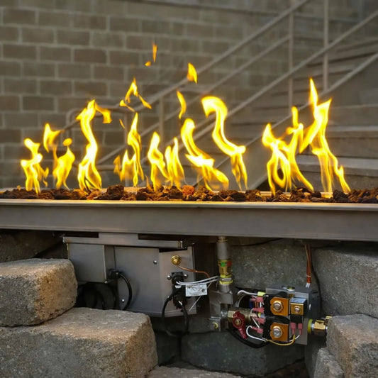Close-up lifestyle image of the HPC Fire Hearth Products Controls Torpedo linear trough pan with T burner, showing bright yellow gas flames dancing over decorative lava rock media. The drop-in electronic ignition system with on off hi lo low controls is visible beneath the pan, including gas valves, wiring, and control box, installed within a stone fire feature against a brick wall backdrop.