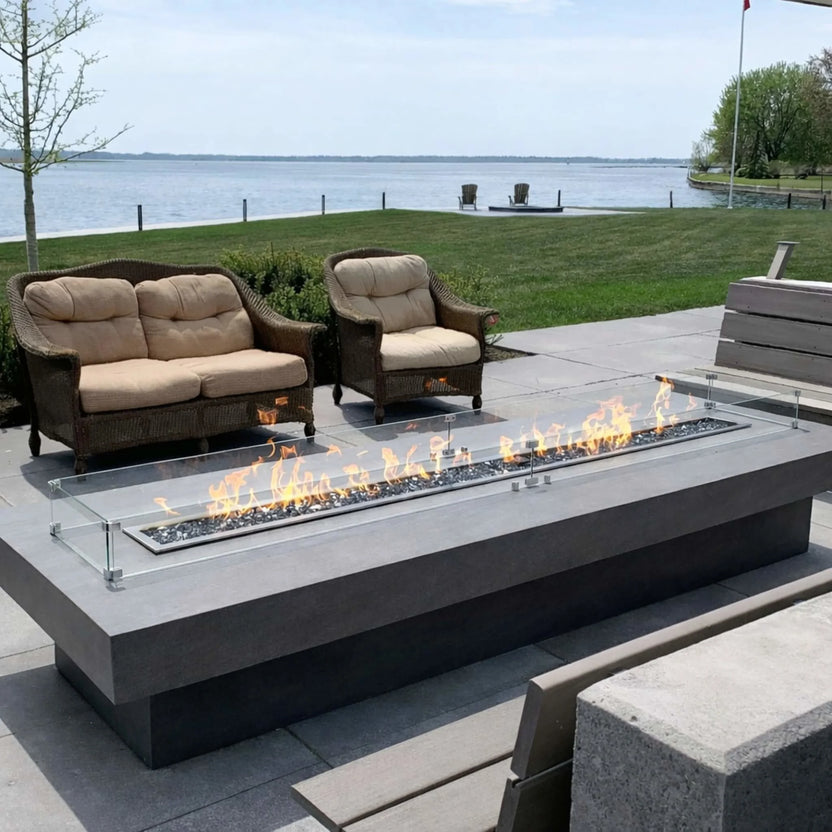 Modern lakeside patio featuring the HPC Torpedo Linear Trough Pan and T Burner fire table with glass wind guard, producing a long, even flame line, surrounded by cushioned wicker seating and overlooking a calm waterfront view.