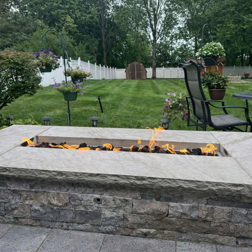 Backyard landscape featuring a stone fire wall with the HPC Torpedo Linear Trough Pan and T Burner, showing a narrow flame strip over lava rock, surrounded by a manicured lawn, white fence, and hanging flower baskets.