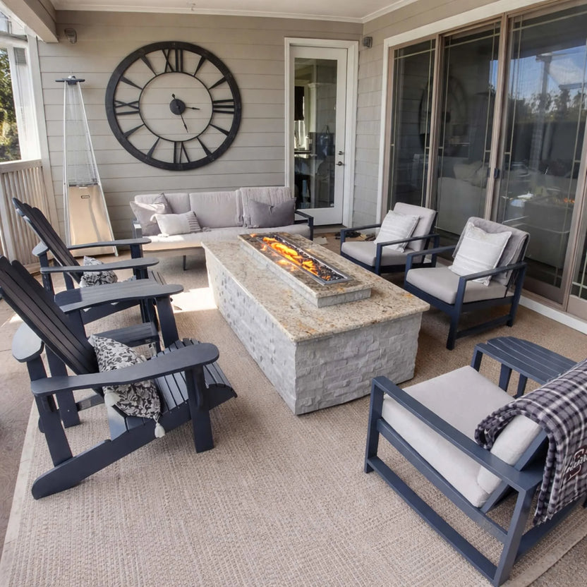 Covered patio seating area with a central stone fire table featuring the HPC Torpedo Linear Trough Pan and T Burner, surrounded by cushioned chairs, Adirondack seating, and a large decorative wall clock for a cozy outdoor lounge.