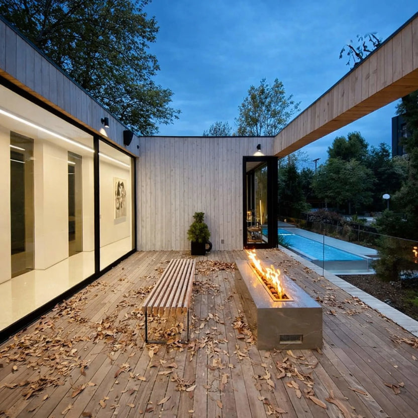 Modern architectural backyard with a sleek linear fire feature using the HPC Torpedo Linear Trough Pan and T Burner, set into a long concrete bench on a wood deck, with a pool and contemporary home design at dusk.