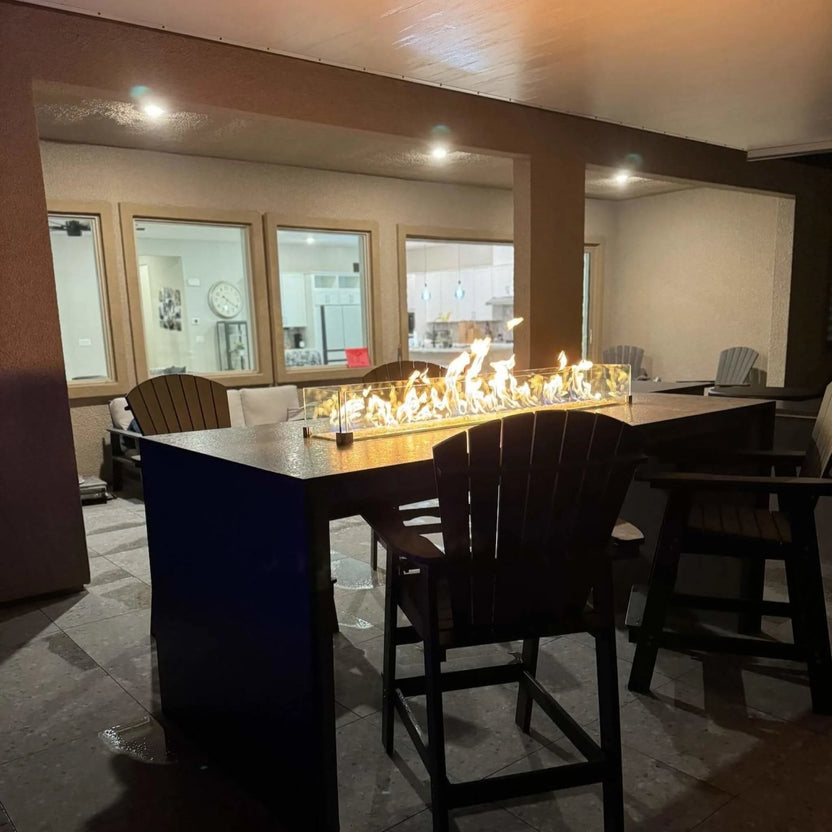 Evening patio bar scene featuring the HPC Torpedo Linear Trough Pan and T Burner integrated into a high-top fire table with glass wind guard, illuminating surrounding seating and creating a warm, inviting outdoor entertainment area.