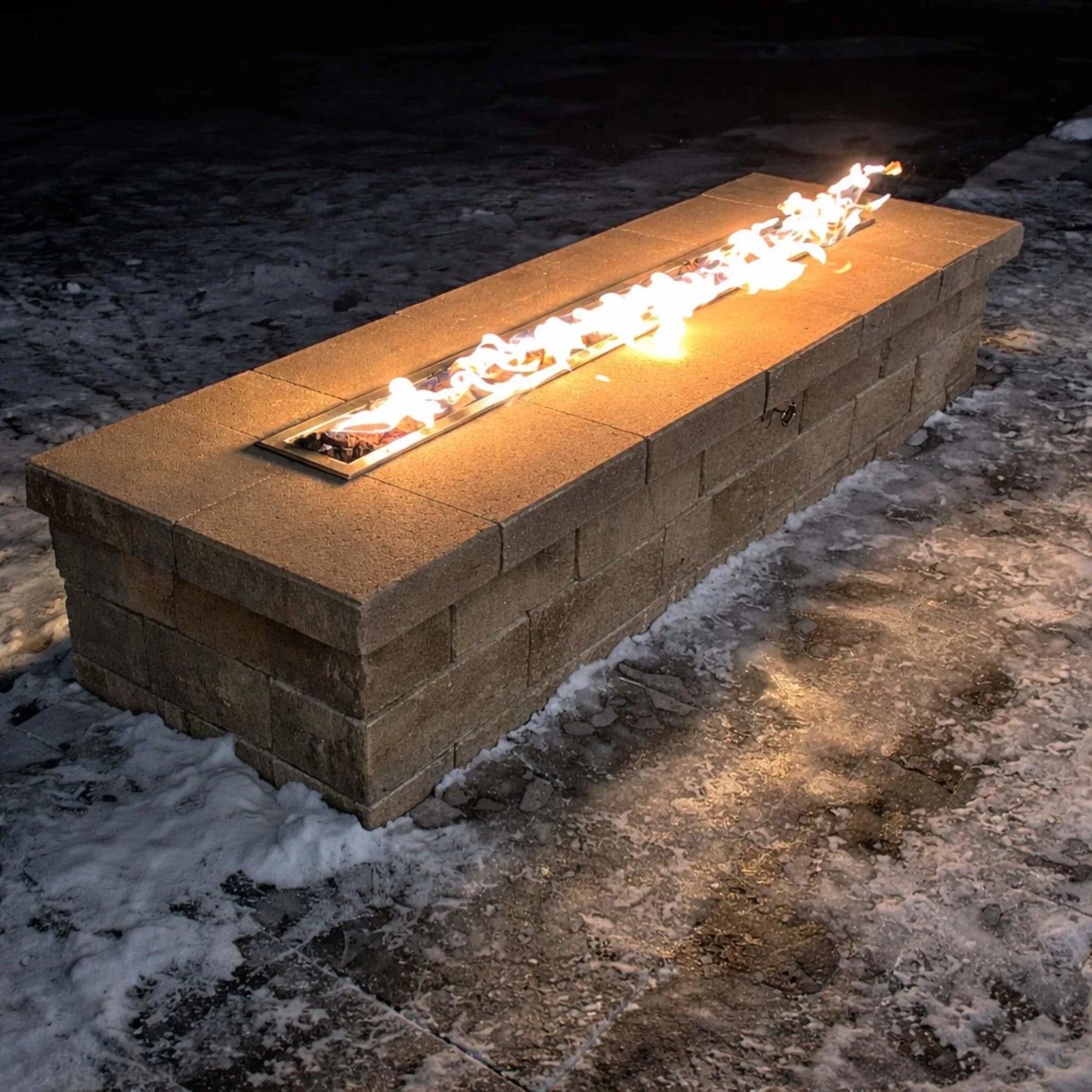 Outdoor winter scene showing the HPC Torpedo Linear Trough Pan and T Burner fire feature embedded in a block-style fire table, with bright flames contrasting against snow-covered ground, demonstrating reliable performance in cold weather.