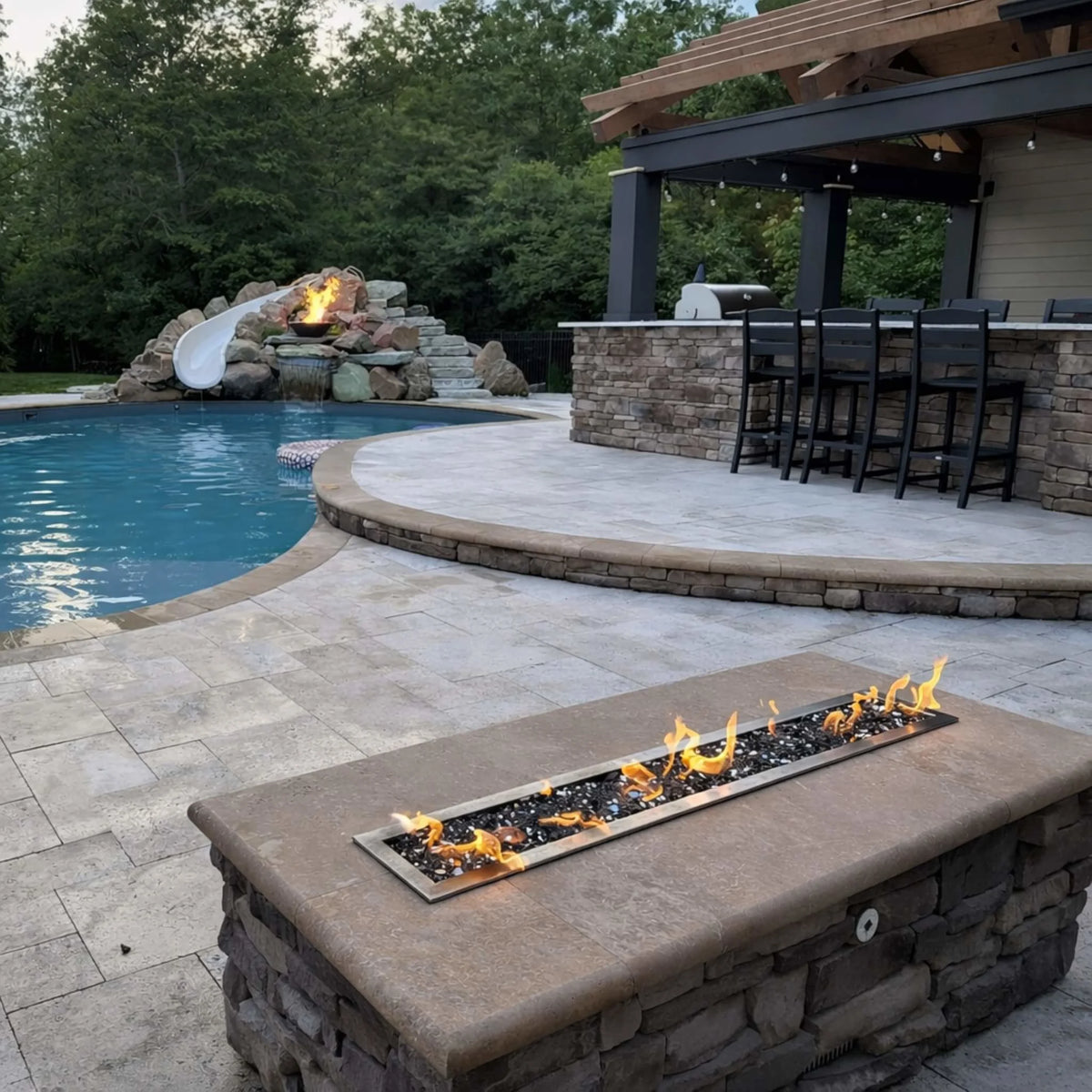 Backyard poolside setup featuring the HPC Torpedo Linear Trough Pan and T Burner in a stone fire table, paired with a pergola bar and outdoor kitchen, highlighting a cohesive luxury outdoor living design with fire and water elements.
