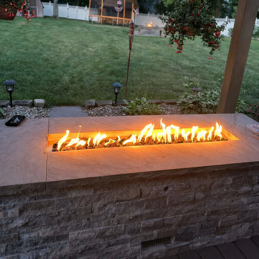 Evening patio scene with the HPC Torpedo Linear Trough Pan and T Burner glowing inside a stone fire feature, casting warm light across a landscaped backyard with lawn, hanging plants, and decorative lighting.