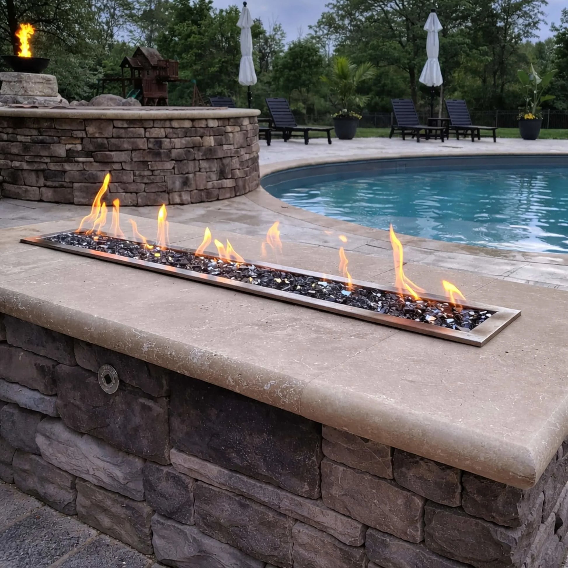 Poolside fire feature using the HPC Torpedo Linear Trough Pan and T Burner installed in a curved stone structure, with a long linear flame bed surrounded by black fire glass, complementing the outdoor pool and lounge area.