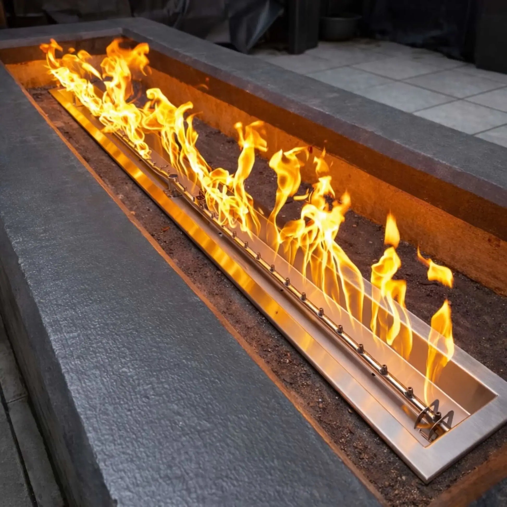 Modern rectangular concrete fire pit featuring a stainless steel linear burner pan with tall yellow flames dancing across the length. The sleek installation is set into a contemporary outdoor seating area, showcasing a clean, built-in design ideal for luxury patios and backyard entertaining spaces.