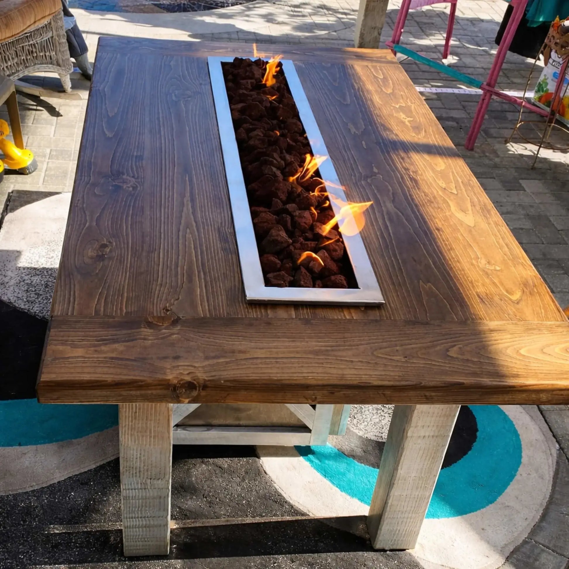 Rustic wood fire table with a built-in stainless steel linear burner filled with lava rock, producing natural yellow flames. The handcrafted wooden top and sturdy legs give it a farmhouse feel, while the sleek burner insert adds a modern touch, making it a functional and stylish outdoor fire feature.