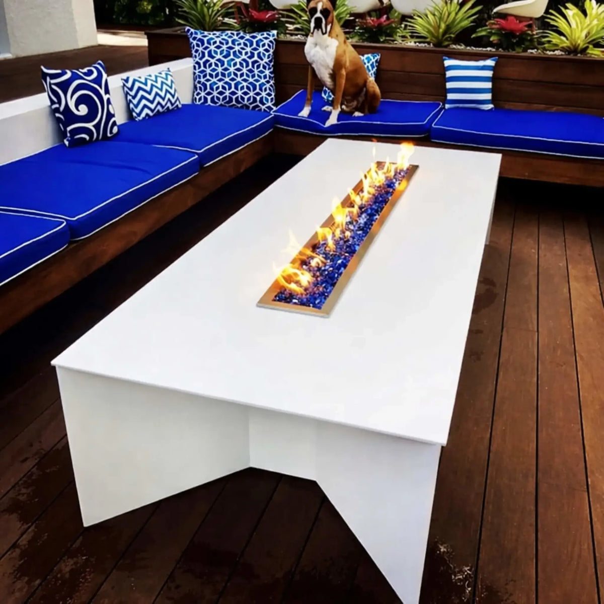 Modern outdoor seating area with a white rectangular fire table showcasing the HPC Fire Hearth Products Torpedo Linear Trough Pan and T Burner Match Lit CSA MLFPK FLEX system. Vibrant blue fire glass enhances the clean linear flame pattern. This CSA match lit fire pit burner offers straightforward lighting, consistent performance, and a stylish focal point for contemporary patio spaces.
