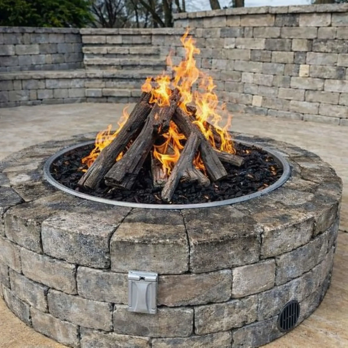 Outdoor circular stone fire pit filled with black lava rock and stacked driftwood-style gas logs burning with tall, vibrant flames. Surrounded by a gray block retaining wall and patio seating area, the setup showcases a natural, rustic fire feature ideal for backyard entertaining and landscape design.