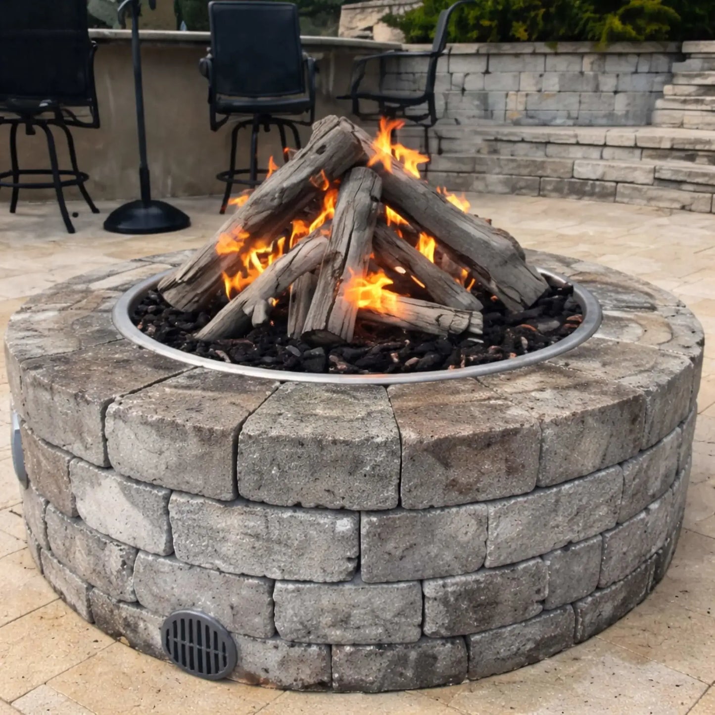 Close-up view of a round stone gas fire pit with Western driftwood logs arranged in a teepee-style stack over lava rock. Flames flicker through the logs, creating a warm ambiance on a paved patio with bar stools and seating in the background, highlighting a cozy outdoor living space design.