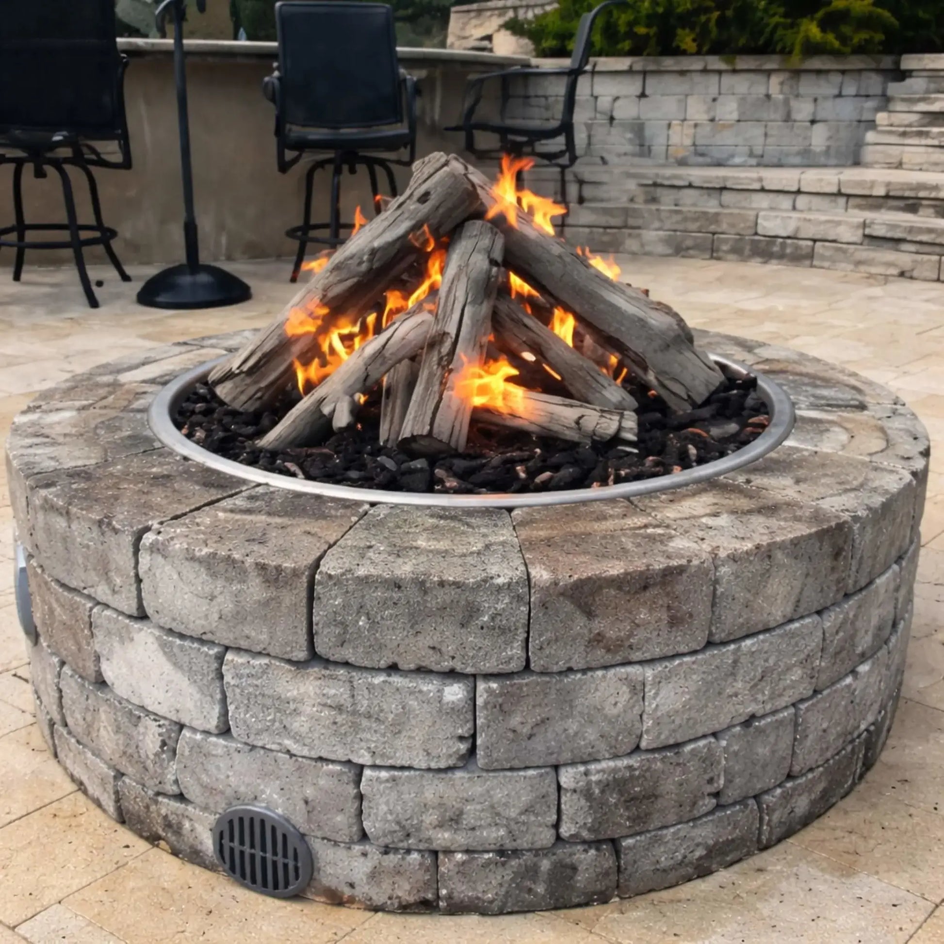 Close-up view of a round stone gas fire pit with Western driftwood logs arranged in a teepee-style stack over lava rock. Flames flicker through the logs, creating a warm ambiance on a paved patio with bar stools and seating in the background, highlighting a cozy outdoor living space design.