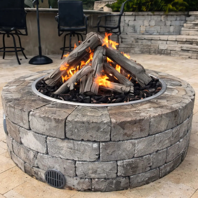 Close-up view of a round stone gas fire pit with Western driftwood logs arranged in a teepee-style stack over lava rock. Flames flicker through the logs, creating a warm ambiance on a paved patio with bar stools and seating in the background, highlighting a cozy outdoor living space design.