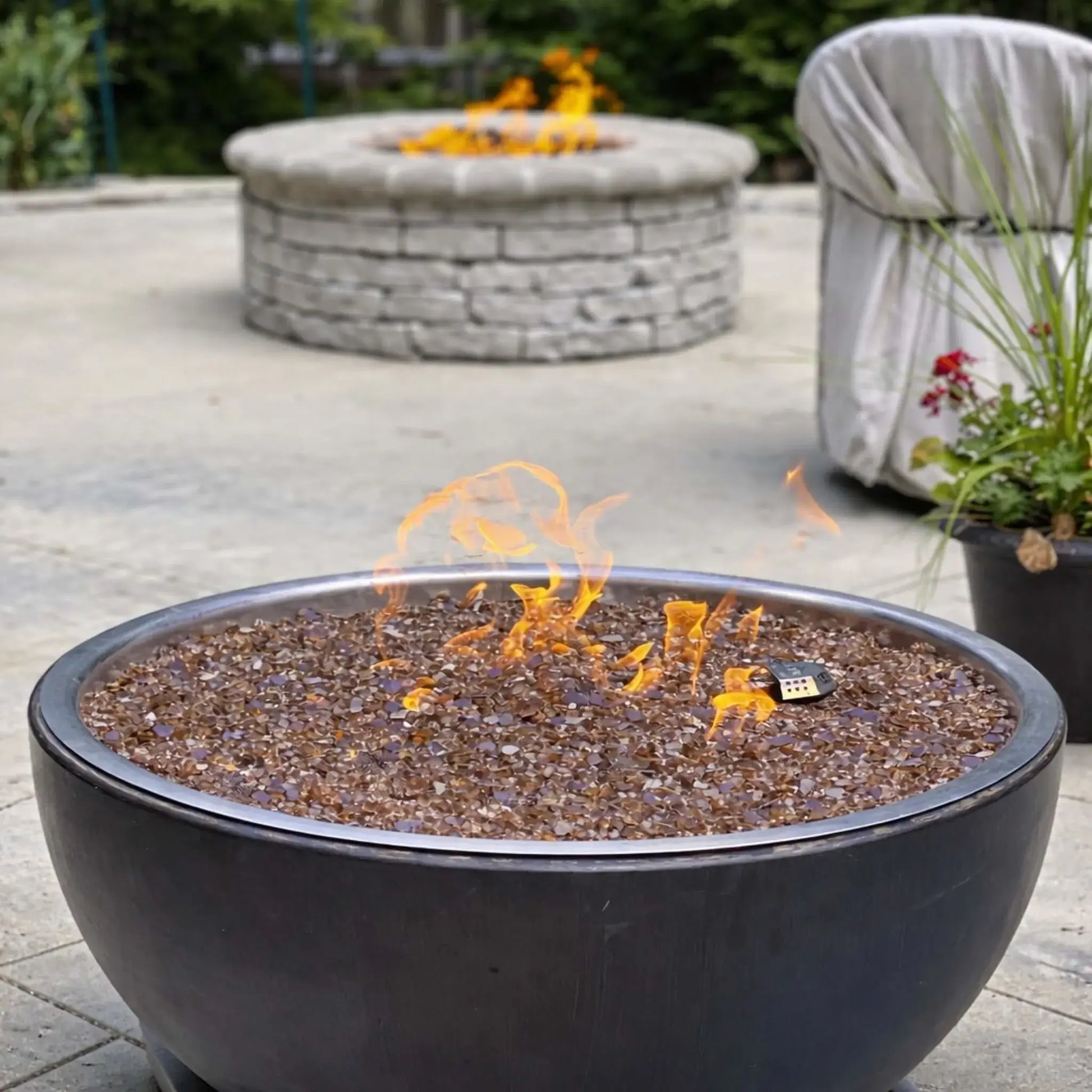 HPC Fire Inspired Hearth Products walnut penta torpedo fire bowl in a backyard patio setting, filled with amber fire glass and lit flames, placed on stone pavers with outdoor seating nearby, creating a cozy residential fire feature.