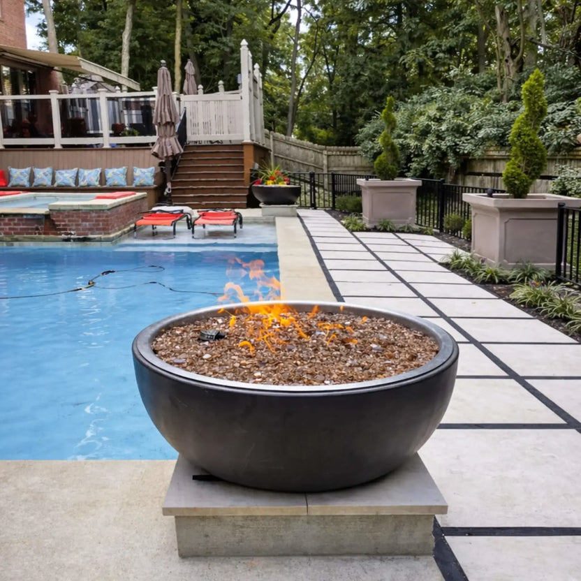 HPC Fire Inspired Hearth Products walnut penta torpedo fire bowl positioned beside a swimming pool, featuring glowing flames over fire glass, surrounded by a landscaped patio, lounge chairs, and modern outdoor living elements.