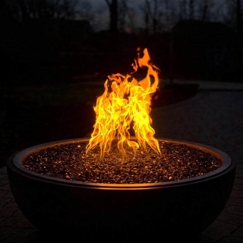 HPC Fire Inspired Hearth Products walnut penta torpedo fire bowl at night with bright flames rising above dark fire glass, creating a dramatic outdoor ambiance and highlighting the fire pit’s warm glow in a low-light setting.