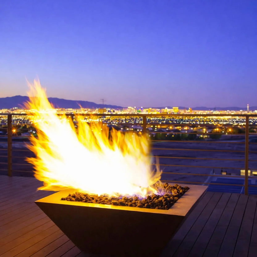 Lifestyle image of a Sedona 40 x 40 Square Series copper fire bowl from HPC Fire Inspired, featuring a powerful flame rising from black fire media on a modern rooftop deck. The fire pit overlooks a glowing city skyline at dusk, with mountains in the background and sleek metal railing, highlighting the dramatic flame output and upscale outdoor living ambiance.