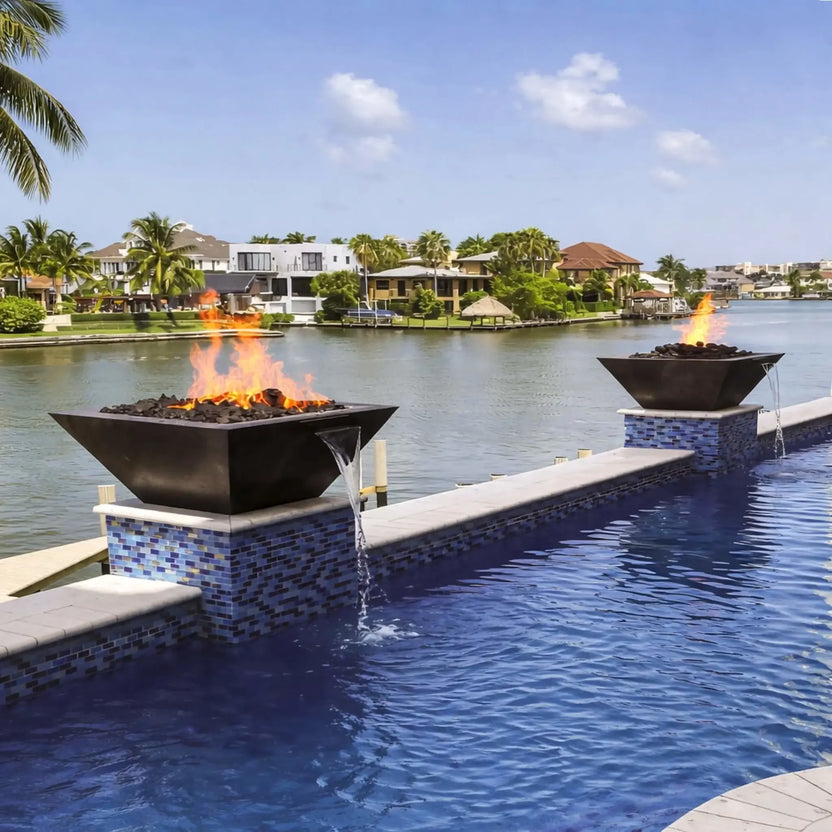 Outdoor luxury pool design showcasing HPC Fire Inspired Sierra Series 36 x 36 inch FireWater bowl with integrated water scupper. Flames rise above lava rock while water cascades into the pool below, mounted on blue tile pedestals along a waterfront canal with tropical landscaping and upscale homes.