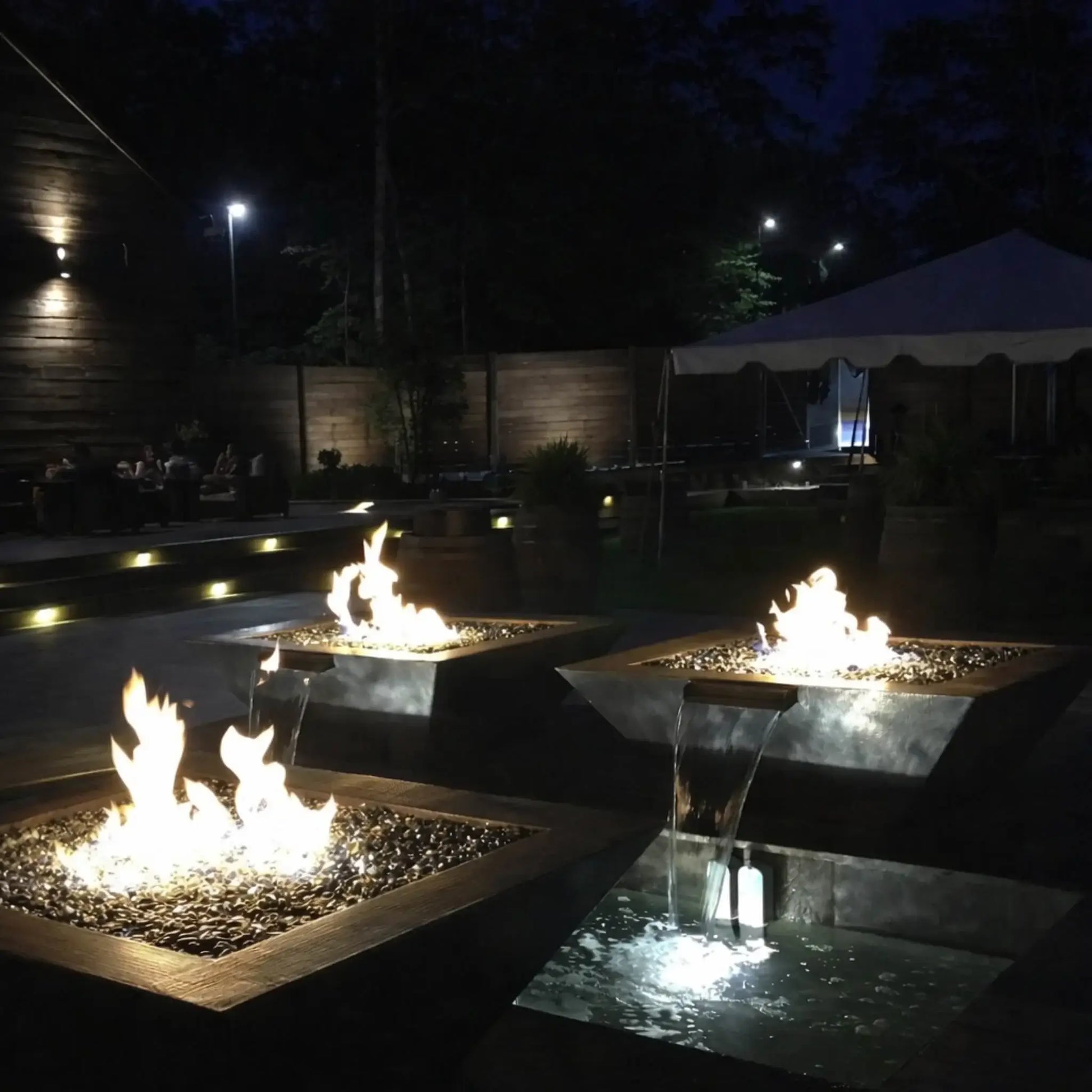Evening outdoor setting featuring HPC Fire Inspired Sierra Series 36 x 36 inch FireWaterbowl with integrated water scupper, where bright flames rise above dark fire glass while water gently cascades into illuminated basins below. Multiple square fire and water bowls create a warm, upscale ambiance in a modern patio space with soft landscape lighting and seating areas in the background.