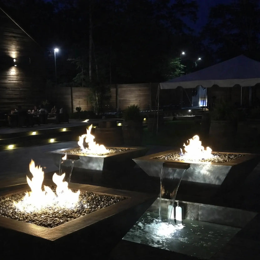 Evening outdoor setting featuring HPC Fire Inspired Sierra Series 36 x 36 inch FireWaterbowl with integrated water scupper, where bright flames rise above dark fire glass while water gently cascades into illuminated basins below. Multiple square fire and water bowls create a warm, upscale ambiance in a modern patio space with soft landscape lighting and seating areas in the background.