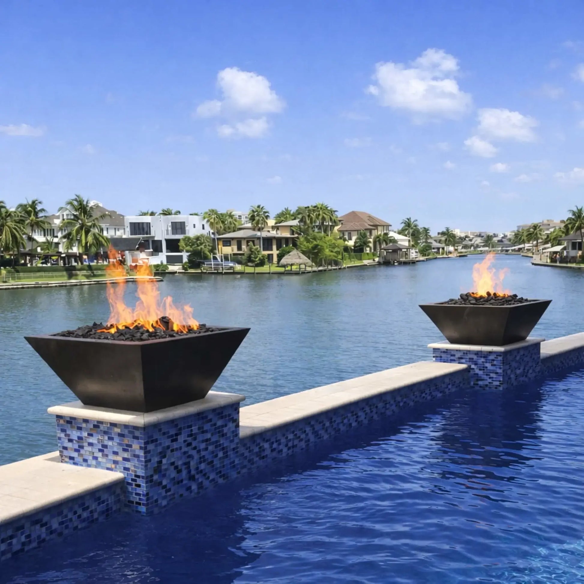 Luxury waterfront poolside installation featuring HPC Fire Inspired Sierra Series 36 x 36 inch fire bowl with black finish, filled with lava rock and active flame. Dual fire bowls sit on tiled pedestals overlooking a canal with modern homes and palm trees, creating a high-end outdoor fire feature ambiance.