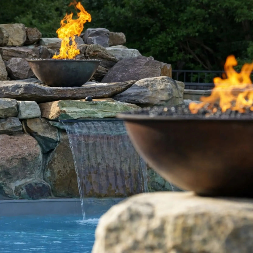 Close-up lifestyle view of HPC Fire Inspired Tempe Series copper fire bowl set on natural stacked stone beside a pool waterfall. Bright yellow flames rise from dark fire media, while water cascades gently into the pool below. A second blurred fire bowl in the foreground adds depth, highlighting the premium outdoor fire and water feature design.