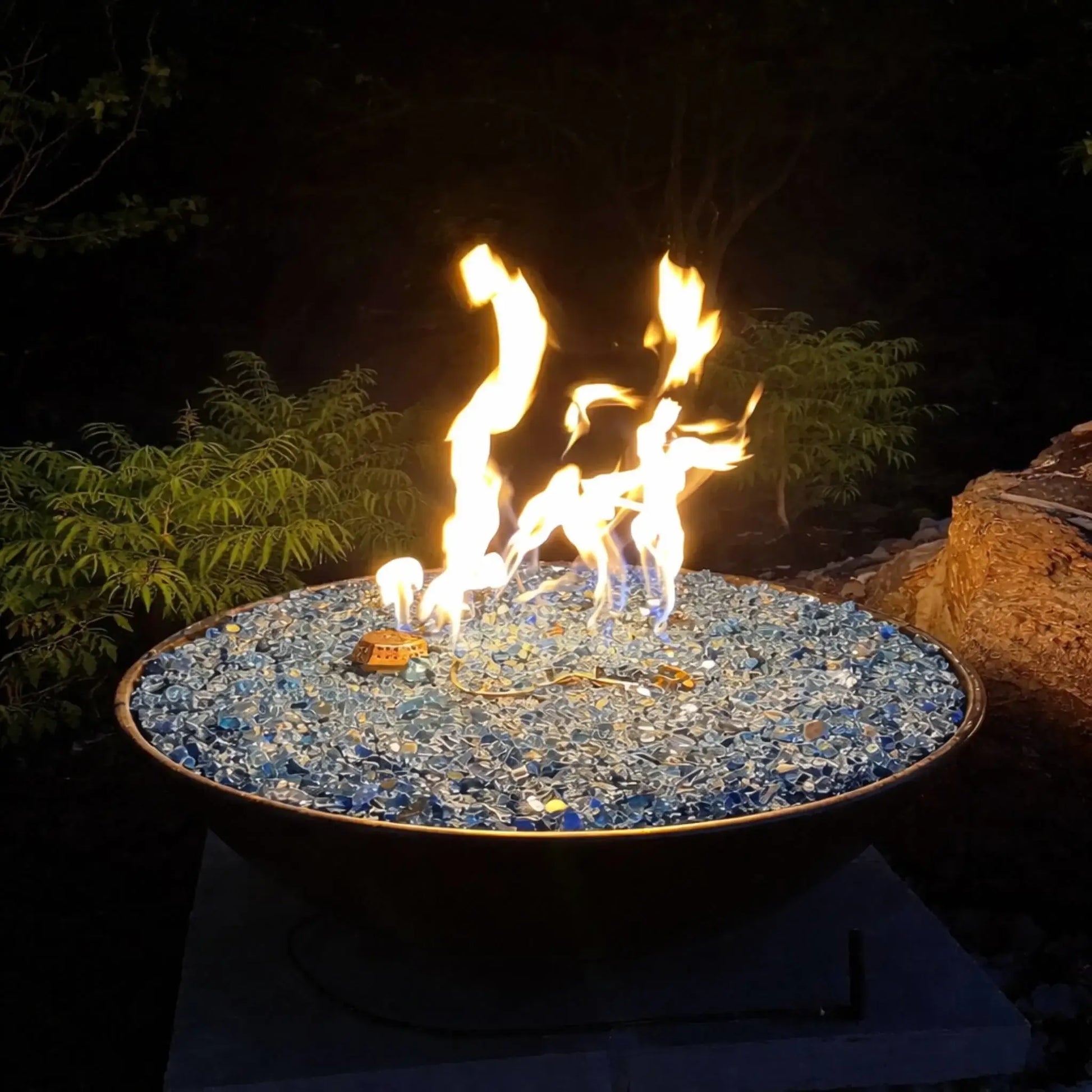 Nighttime close-up of HPC Fire Inspired Tempe Series copper fire bowl filled with reflective blue fire glass, with tall, bright flames dancing against a dark backyard backdrop. The hammered copper rim and glowing fire create a warm focal point, while surrounding foliage and stone elements subtly frame this premium outdoor fire feature.