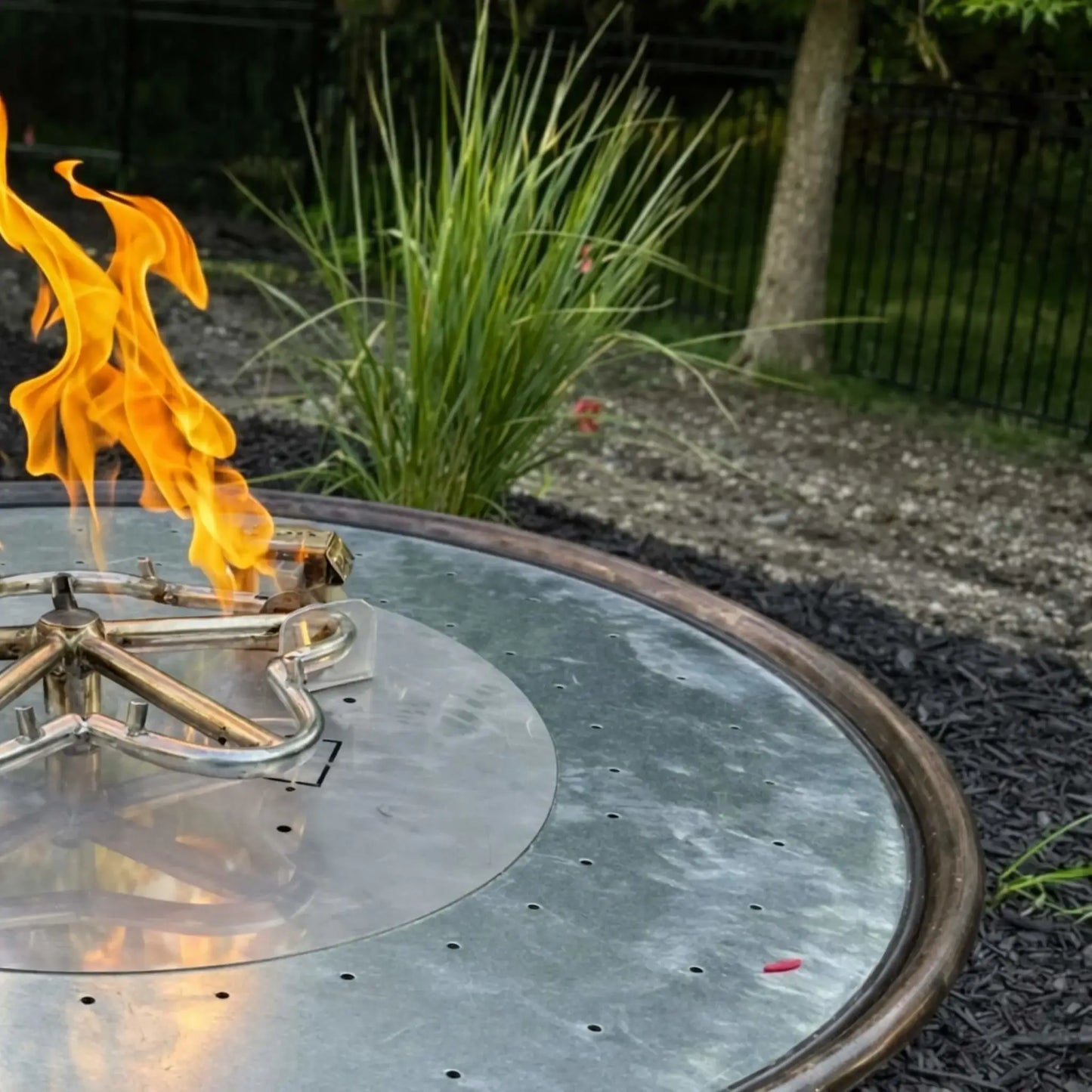 Detailed view of HPC Tempe Series fire bowl burner assembly installed in a copper bowl, showing stainless steel burner ring and ignition components before media is added. A live flame burns at one section, highlighting the internal structure, with landscaped greenery and fencing visible in the background for a real-world installation context.