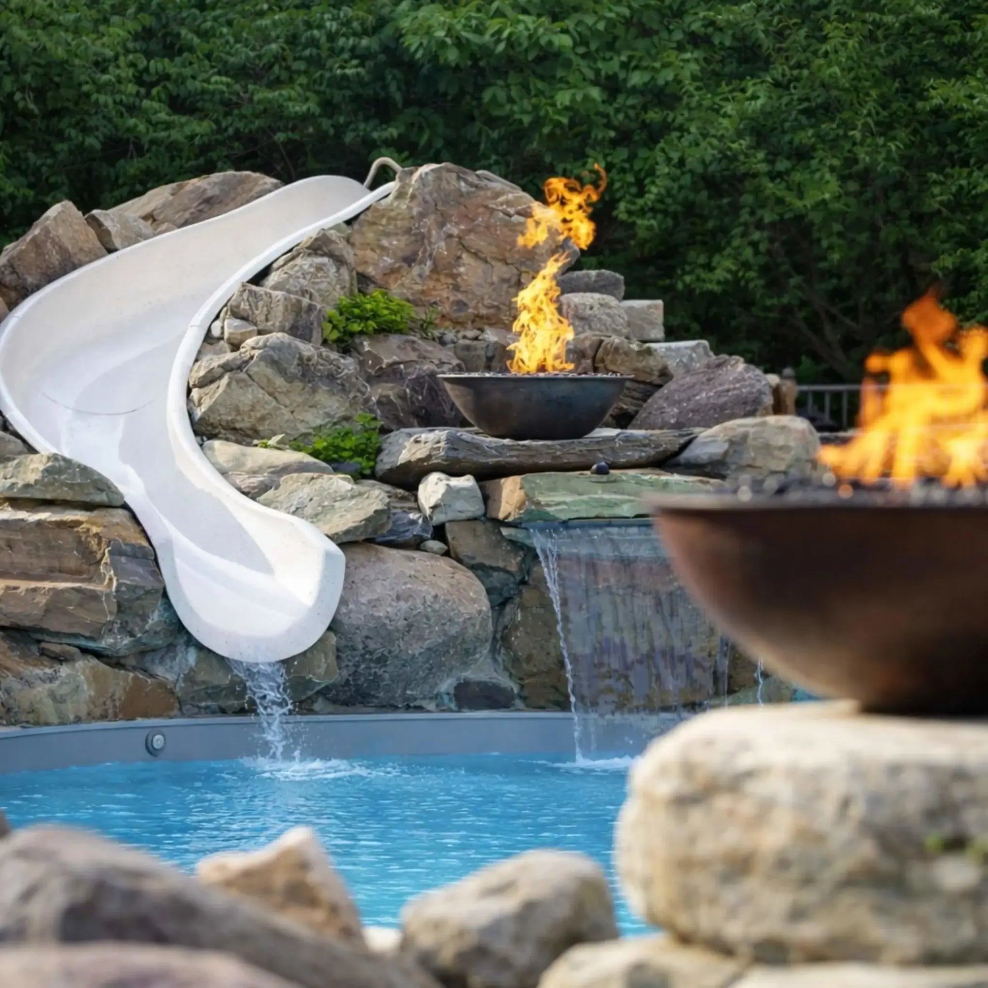 Backyard pool scene featuring HPC Tempe Series copper fire bowl integrated into a rock waterfall landscape with a curved white pool slide. Flames burn steadily above the bowl while water flows beneath into the pool, creating a resort-style setting surrounded by natural stone and lush greenery.