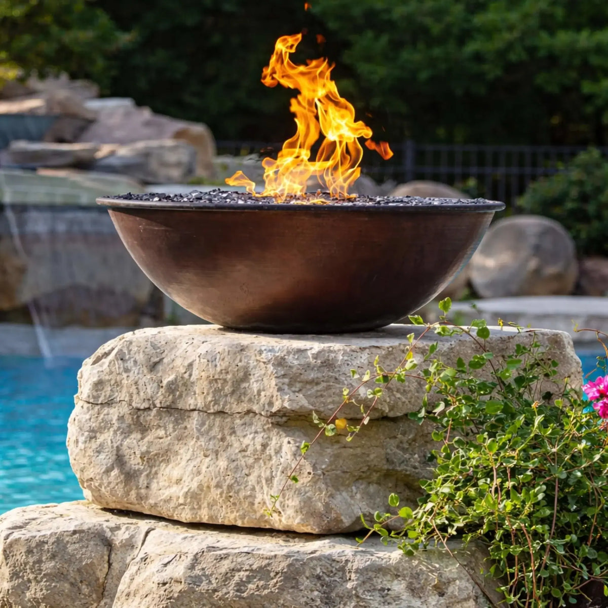 Detailed close-up of an HPC Tempe Series copper fire bowl placed on a stone pedestal near a pool, showcasing rich patina tones and active flames dancing above dark fire media. Background waterfalls and landscaping provide a soft, natural backdrop that emphasizes the bowl’s craftsmanship and outdoor elegance.
