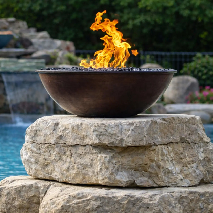 Front-facing view of HPC Tempe Series copper fire bowl elevated on stacked stone blocks beside a pool, surrounded by flowering plants and greenery. Flames flicker above the burner, while a second matching fire bowl and cascading water feature appear softly in the background, reinforcing a cohesive outdoor design.