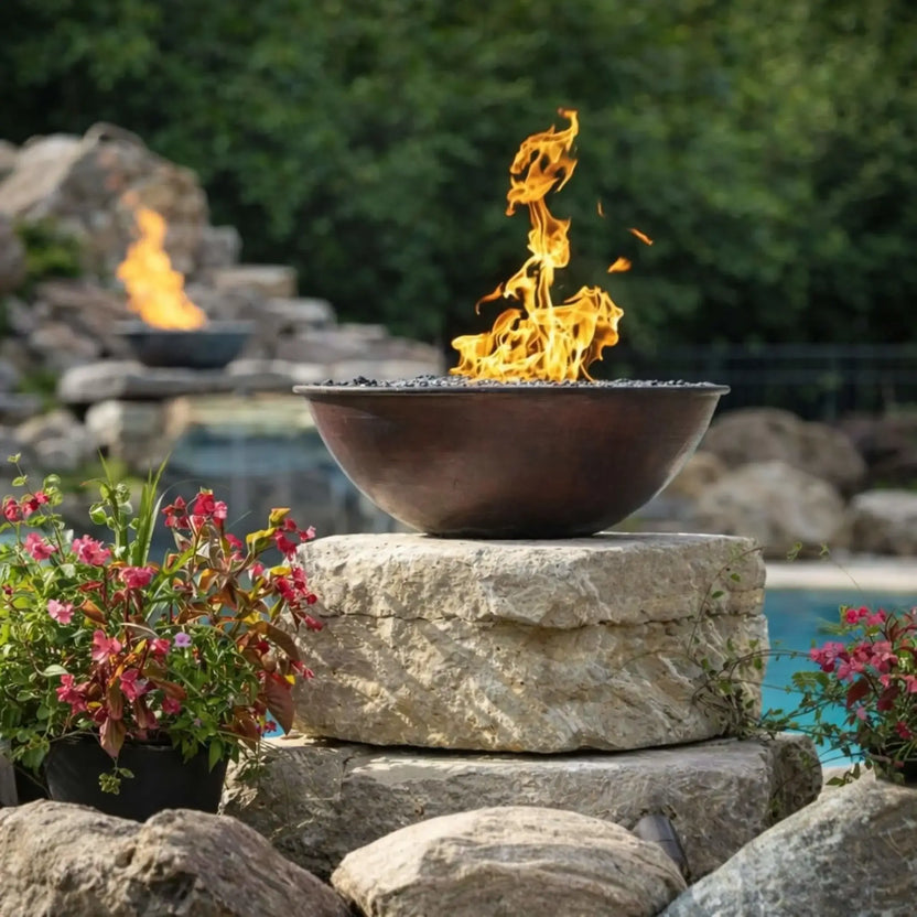 Daytime patio scene featuring HPC Tempe Series fire bowl filled with blue fire glass, positioned on a square pedestal near a swimming pool. Flames dance across the glass surface, reflecting sunlight, with a wooded backyard and stone water feature in the distance creating a serene outdoor living environment.