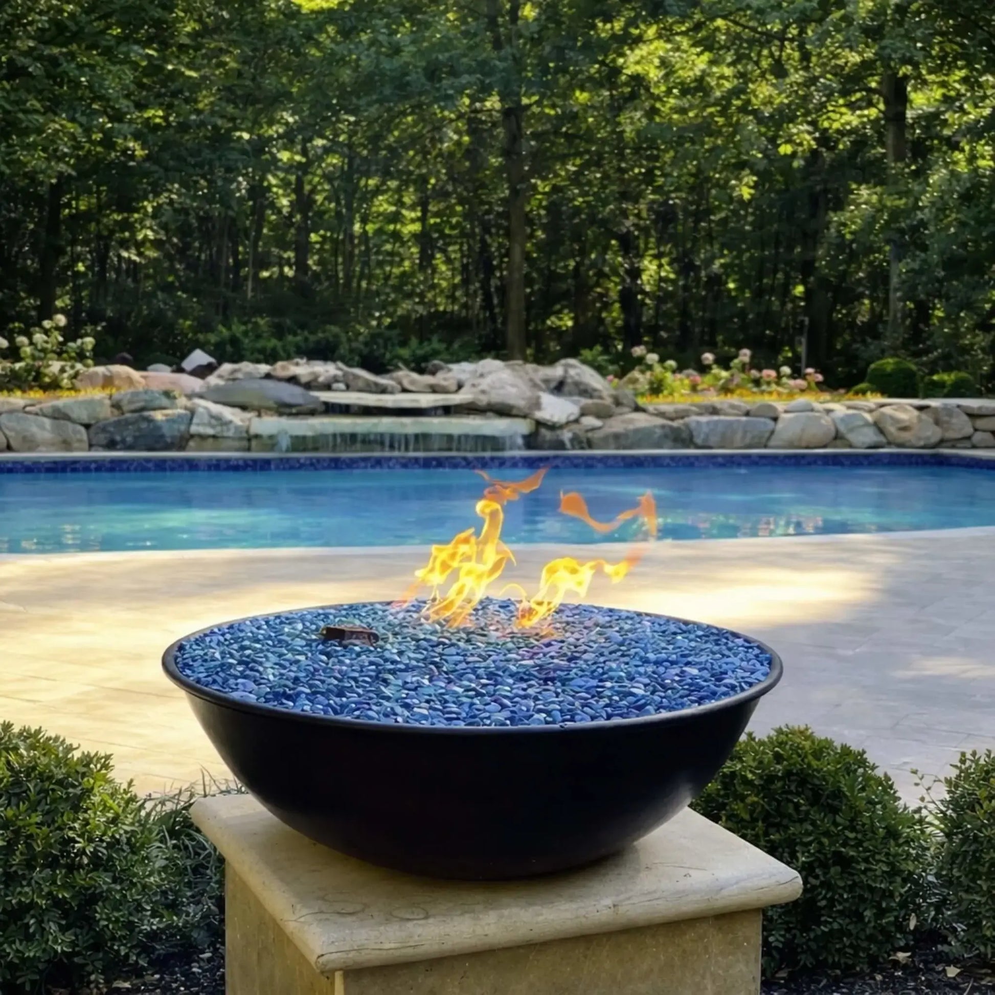 Close-up of HPC Tempe Series fire bowl with vibrant blue fire glass and low, flowing flames, set near a covered patio with columns. The pool and landscaped yard extend into the background, showcasing how the fire feature enhances both modern and traditional outdoor living spaces.