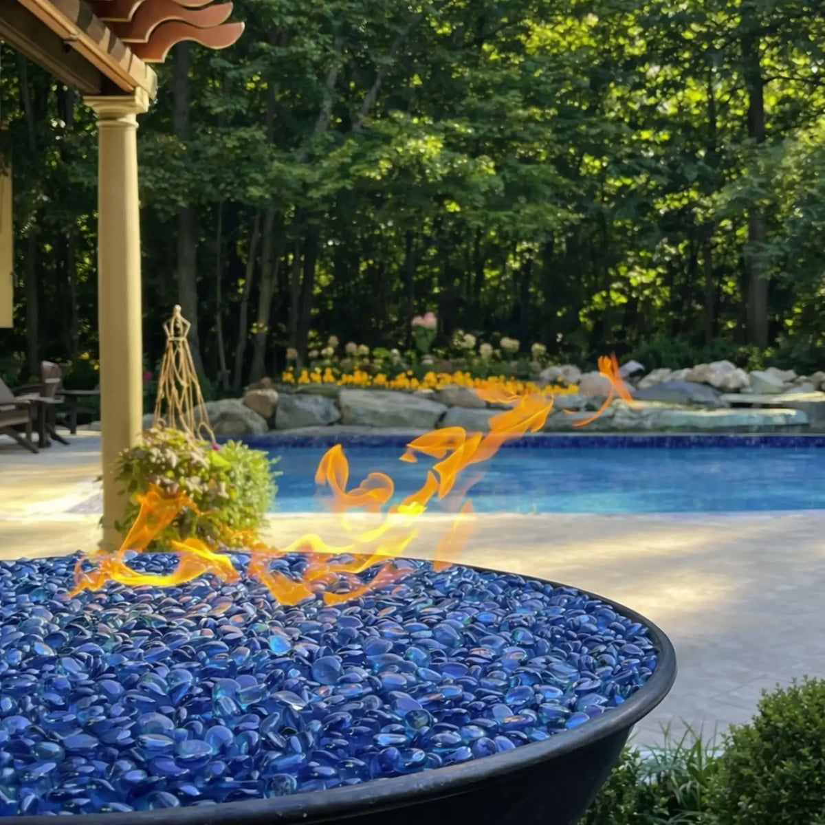 Wide backyard view highlighting multiple HPC Tempe Series copper fire bowls installed around a natural stone poolscape. Flames glow atop each bowl while rock formations, waterfalls, and surrounding trees create a tranquil, resort-style atmosphere ideal for outdoor entertaining.