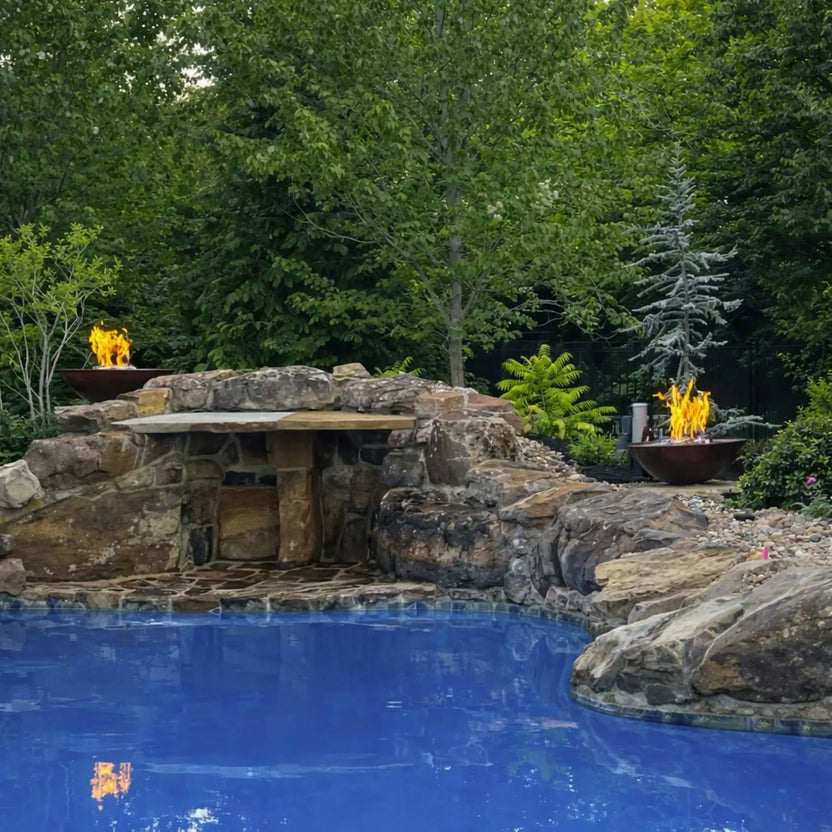Expansive poolside landscape featuring HPC Tempe Series fire bowls integrated into a curved rock formation with cascading water. Flames reflect on the blue pool surface, while dense greenery frames the scene, emphasizing the dramatic combination of fire and water in a luxury backyard setting.