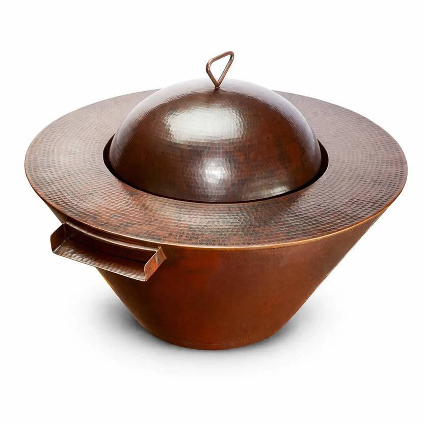 Hammered copper round fire pit burner cover installed on a Mesa-style bowl, featuring a smooth domed lid with a triangular handle and a wide textured rim. The rich reddish-brown patina highlights the handcrafted finish, while the integrated side tray detail adds functionality. Clean white background emphasizes the shape, texture, and premium copper construction.