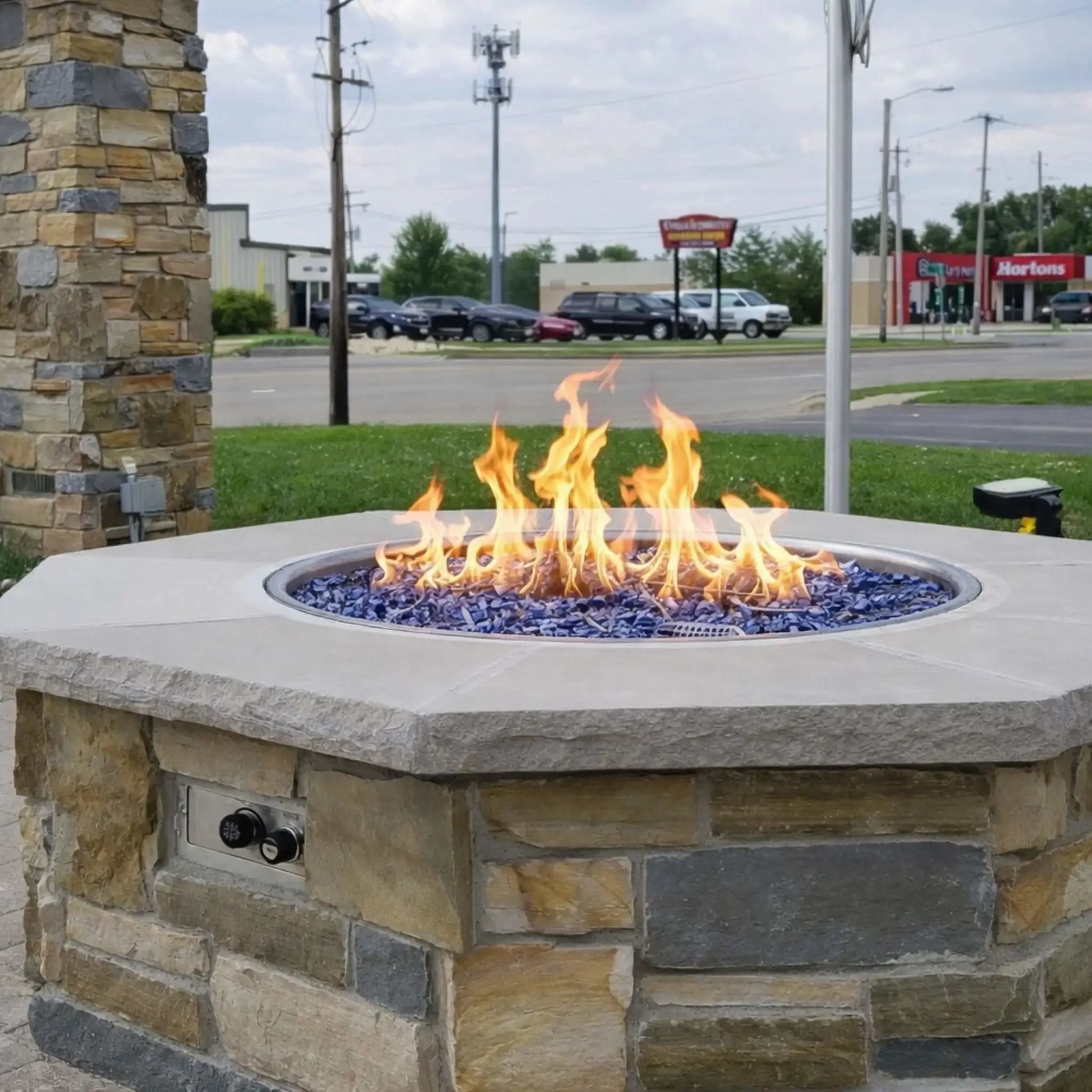 Outdoor octagon fire pit table installed in a commercial patio setting, finished with stacked stone veneer and topped with a smooth concrete cap. A vibrant gas flame burns over blue fire glass media within a round burner pan, while a push-button ignition control panel is visible on the side. The background shows a roadside environment with parked cars and nearby businesses.