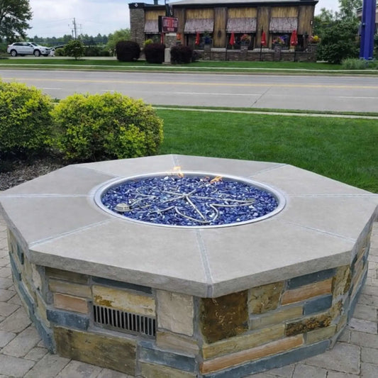 Octagon fire pit table with a finished stone veneer base and light gray concrete top, filled with blue fire glass and a visible stainless steel burner. The unit is installed on a paved outdoor patio area surrounded by manicured grass and landscaping, with a restaurant building and roadway in the background, highlighting a clean, modern commercial outdoor fire feature.