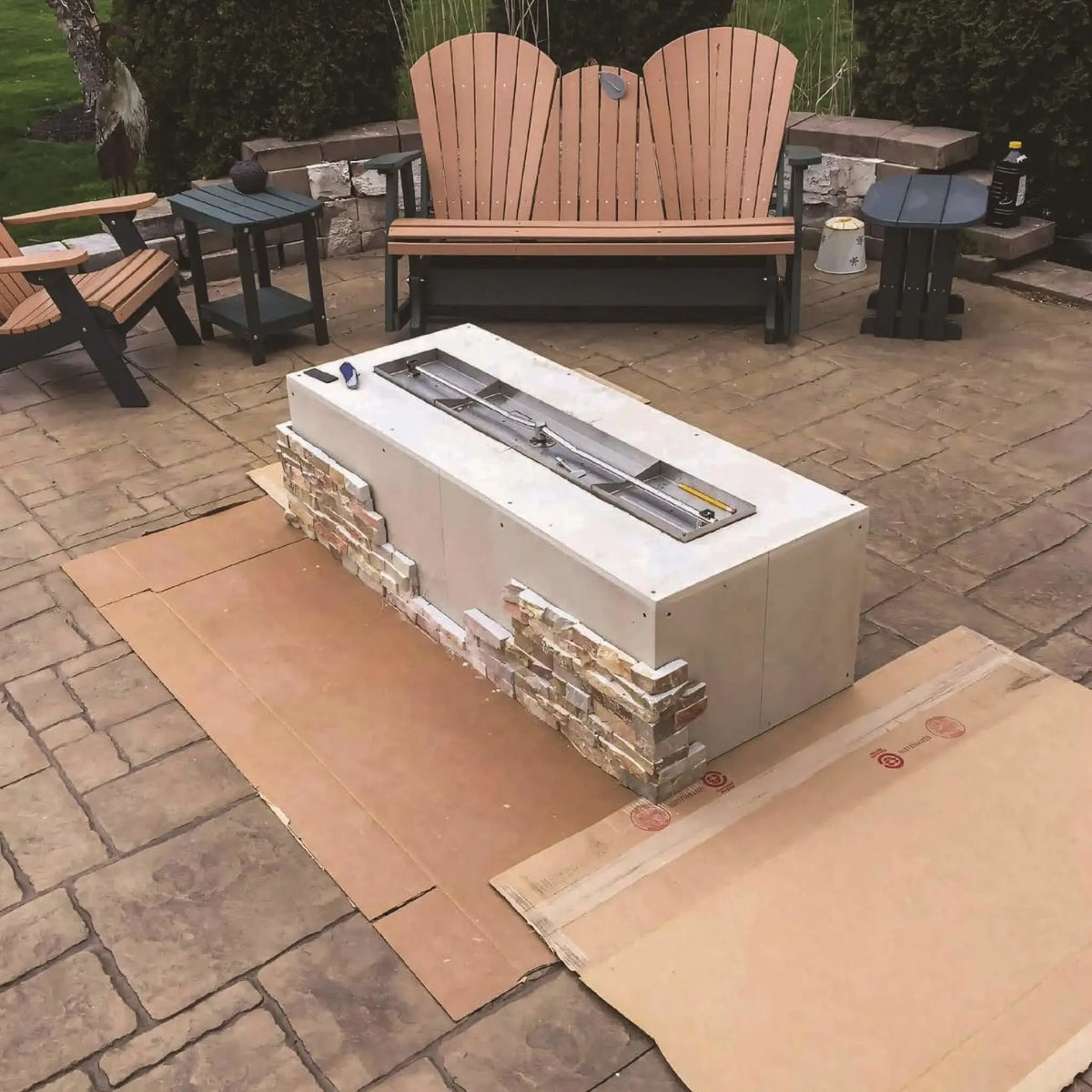 Outdoor patio scene featuring a rectangular ready-to-finish fire pit table under construction, placed on protective cardboard. Surrounded by Adirondack-style chairs and side tables, the linear burner insert is visible, showcasing a modern backyard fire feature installation in progress.