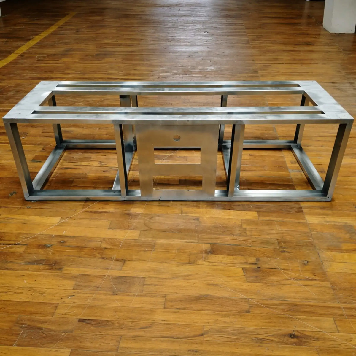 Steel fire pit frame displayed on a wood floor, showing structural support bars and central cutout for burner and control access. The industrial metal finish highlights durability and readiness for custom outdoor fire pit builds.