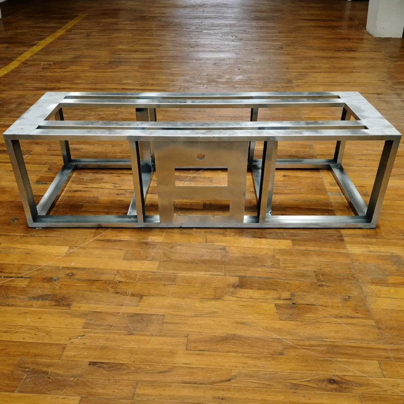 Steel fire pit frame displayed on a wood floor, showing structural support bars and central cutout for burner and control access. The industrial metal finish highlights durability and readiness for custom outdoor fire pit builds.