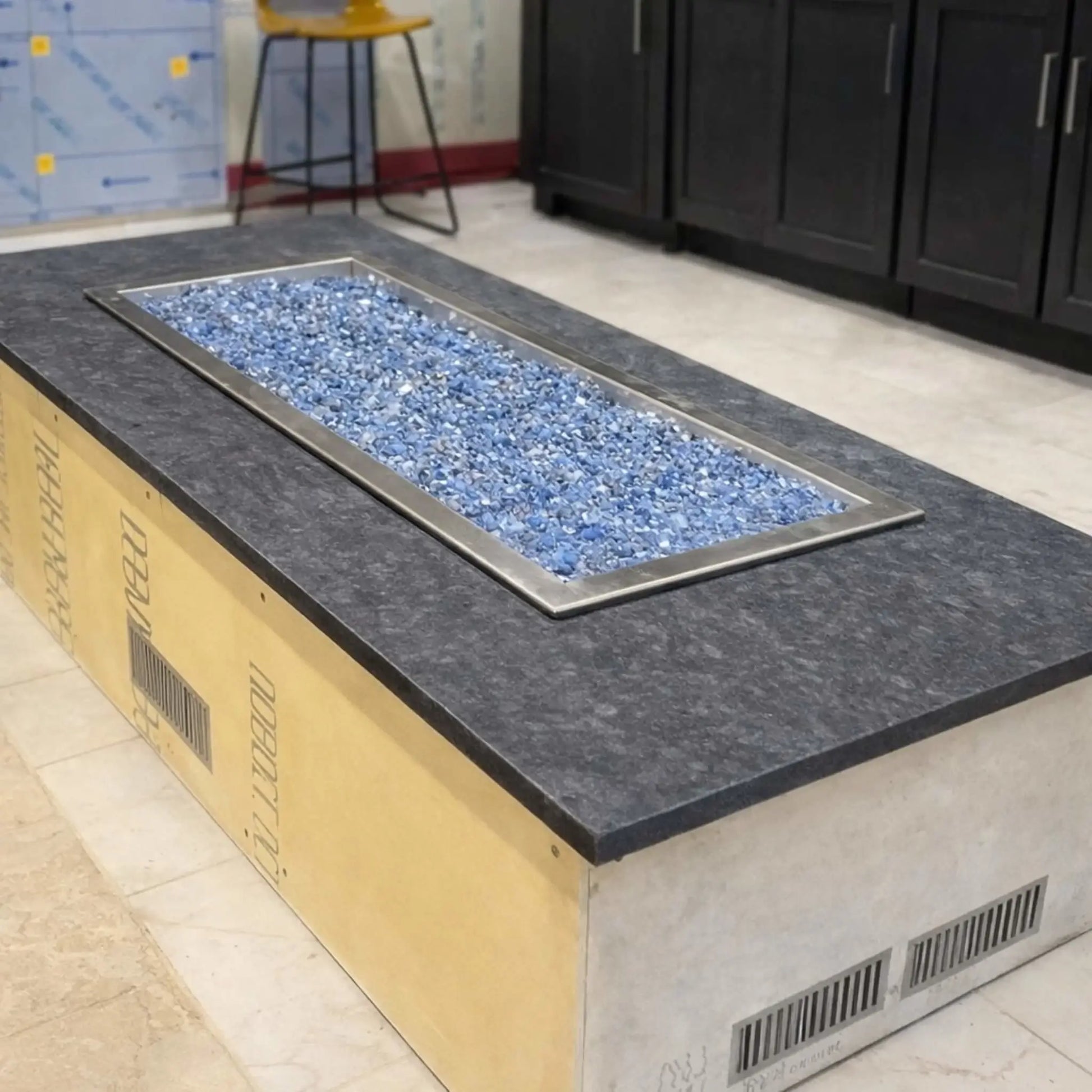 Completed rectangular fire pit table installed indoors with a dark stone top and blue fire glass filling a linear burner. The finished unit includes ventilation slots and showcases a sleek, modern design suitable for custom fire feature installations.