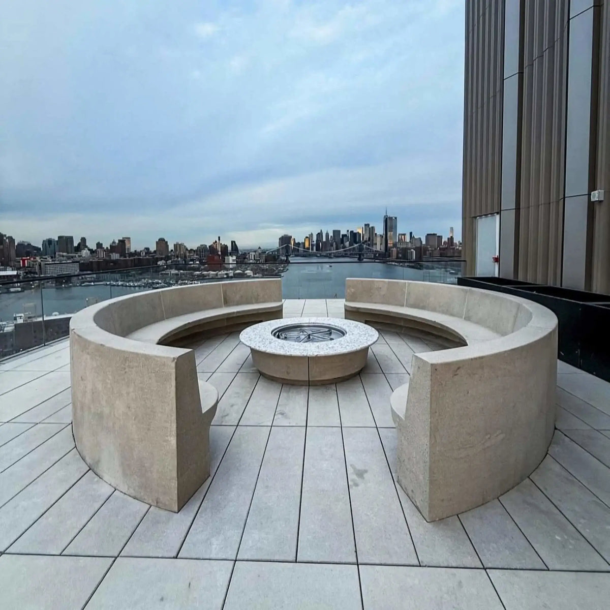 Close-up view of a round stone gas fire pit table with a smooth granite top and black lava rock media, showcasing bright yellow flames at the center. The fire pit is built into a modern backyard patio with cushioned outdoor seating, stone columns, and a manicured lawn bordered by tall pine trees.