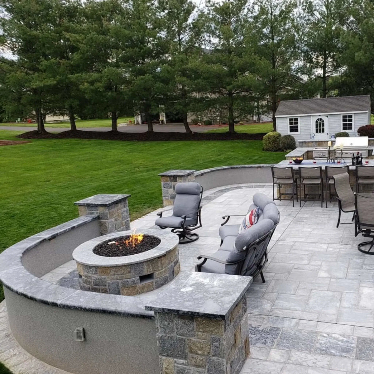 Modern rooftop terrace featuring a circular concrete fire pit table surrounded by curved built-in seating. The fire pit sits on large format pavers and overlooks a city skyline with a river and bridge in the distance, creating a sleek urban outdoor living space with panoramic views.