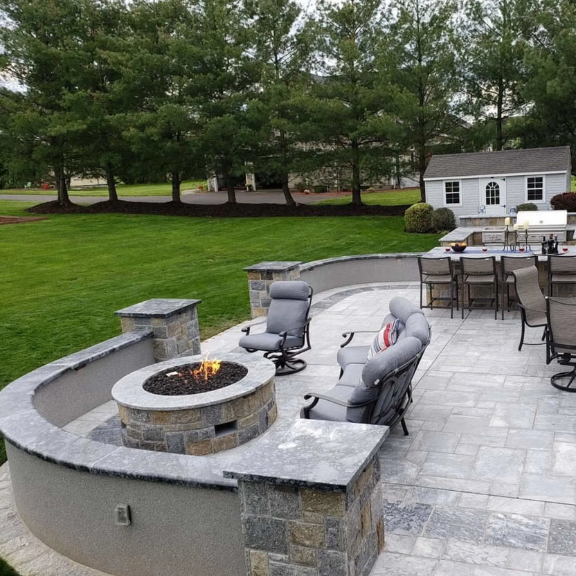 Modern rooftop terrace featuring a circular concrete fire pit table surrounded by curved built-in seating. The fire pit sits on large format pavers and overlooks a city skyline with a river and bridge in the distance, creating a sleek urban outdoor living space with panoramic views.