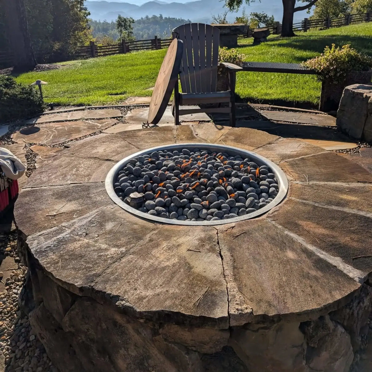 Wide-angle view of a luxury backyard patio with a round stone fire pit table centered in a curved seating wall. The space includes cushioned patio chairs, an outdoor dining area, and a built-in grill station, all set against a large green lawn and mature trees for a spacious outdoor entertaining area.