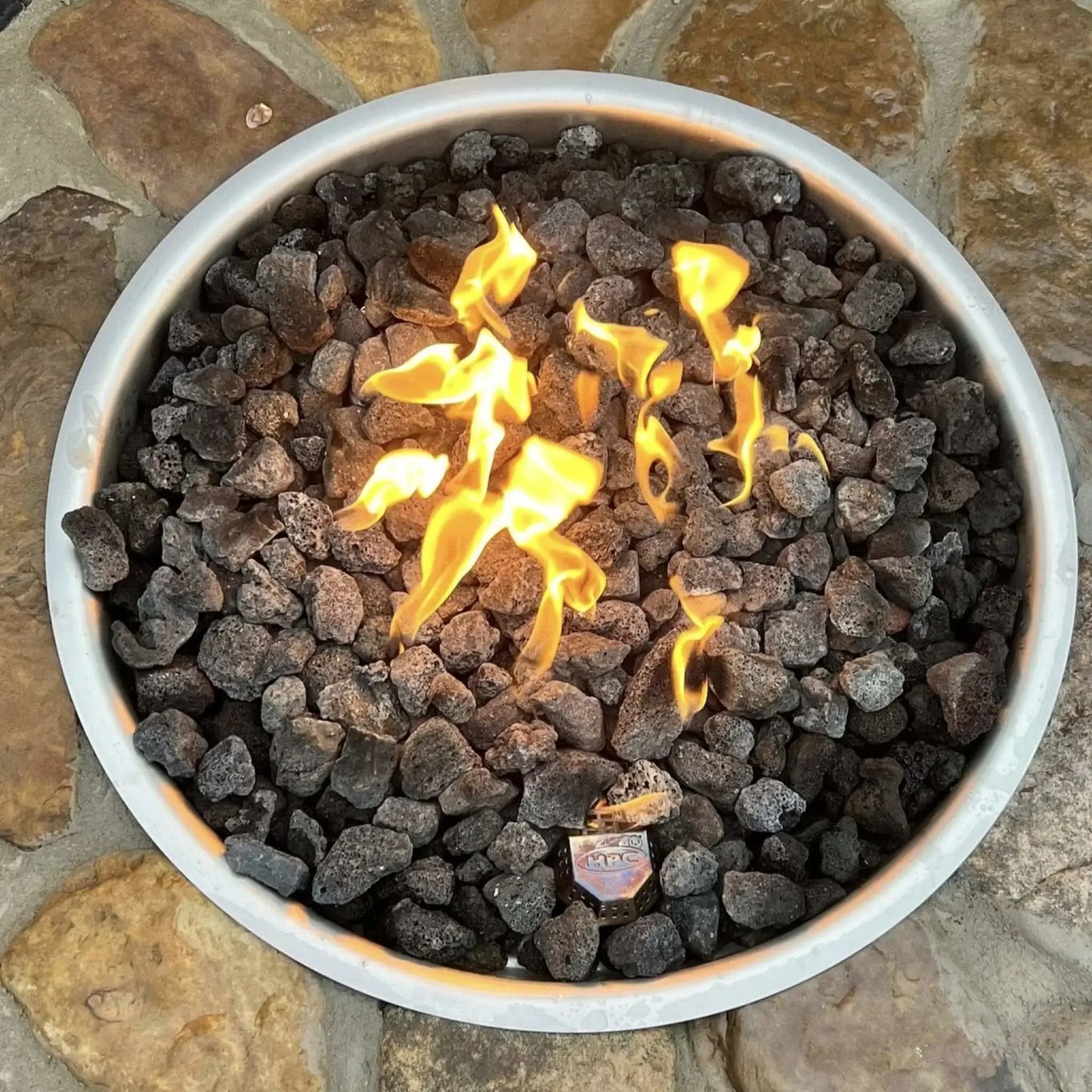 Rustic round fire pit built with natural stone and filled with smooth gray lava rocks, burning with low flames. The fire pit is set into a flagstone patio with scenic mountain views, a wooden Adirondack chair, and a peaceful landscaped yard in the background.