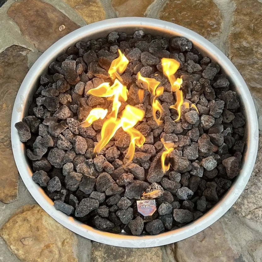Rustic round fire pit built with natural stone and filled with smooth gray lava rocks, burning with low flames. The fire pit is set into a flagstone patio with scenic mountain views, a wooden Adirondack chair, and a peaceful landscaped yard in the background.