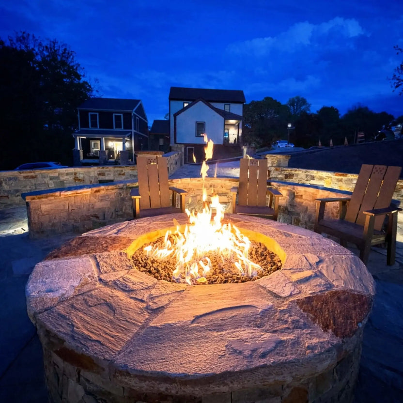 Contemporary circular fire pit table with a smooth concrete top and recessed burner, set in a large paved patio overlooking a landscaped yard with a pond. The clean design emphasizes the burner structure and surrounding stone veneer base in a bright outdoor setting.