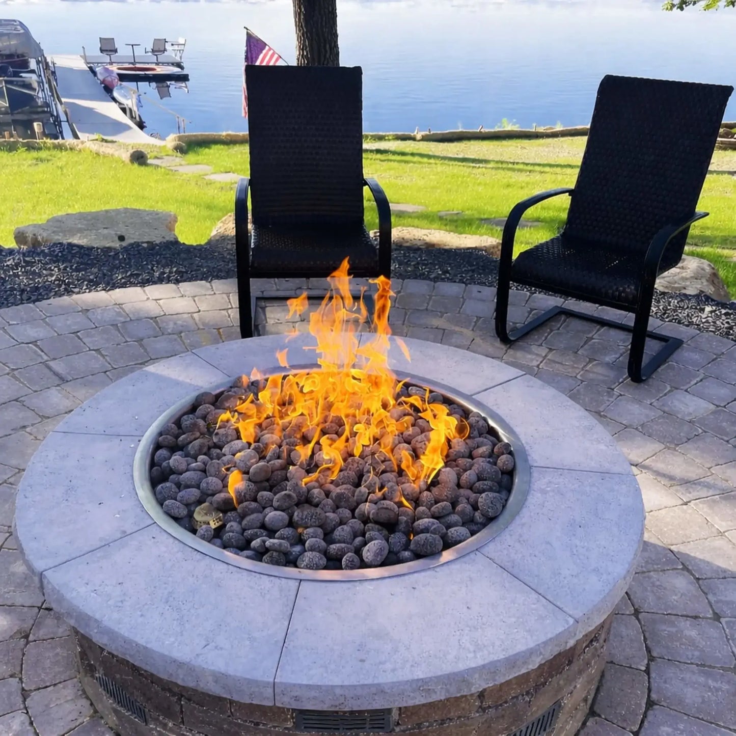Evening backyard scene featuring a round stone fire pit table with active flames illuminating surrounding Adirondack chairs. The fire pit is integrated into a raised stone patio with built-in seating, creating a warm and inviting outdoor gathering space under a deep blue twilight sky.